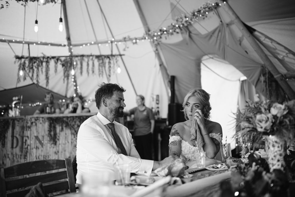 A man and a woman sit at a decorated table inside a marquee; the woman appears emotional while the man holds her hand.