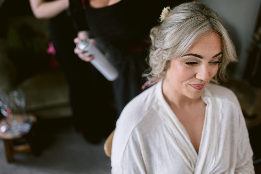 A woman with blonde hair in a white dressing gown sits while another person styles her hair with a can of hairspray in a home setting.