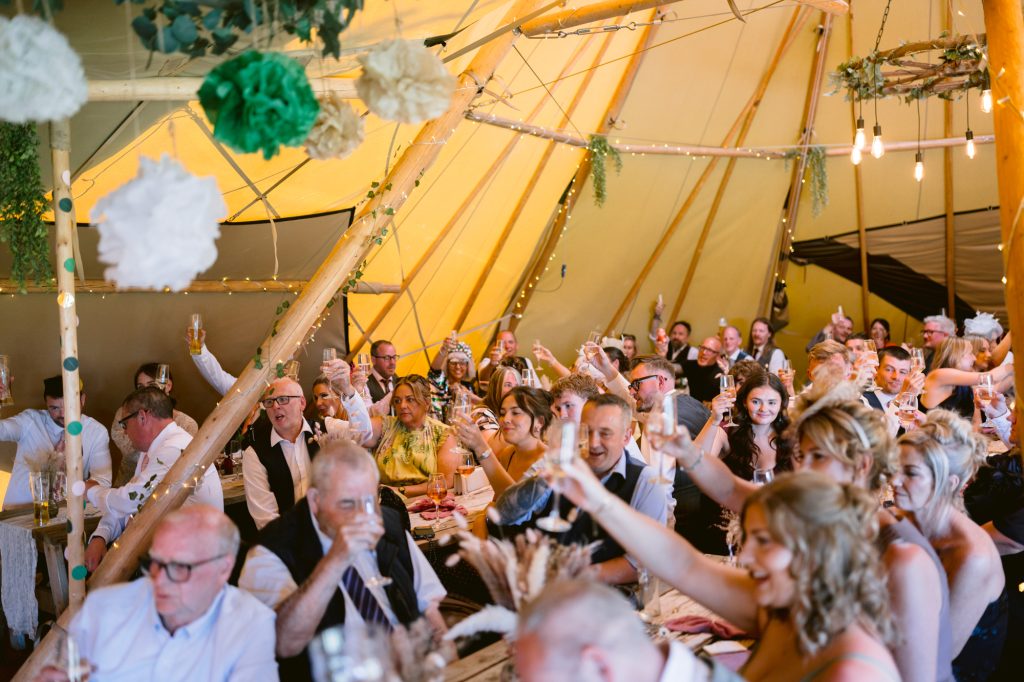 A large group of people seated inside a decorated marquee raise glasses in a celebratory toast during a festive event.