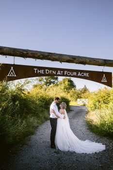A bride and groom embrace on a gravel path under a wooden archway sign that reads "The Den At Four Acre," surrounded by greenery on a sunny day.