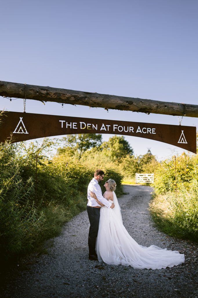 A bride and groom embrace on a gravel path under a wooden archway sign that reads "The Den At Four Acre," surrounded by greenery on a sunny day.