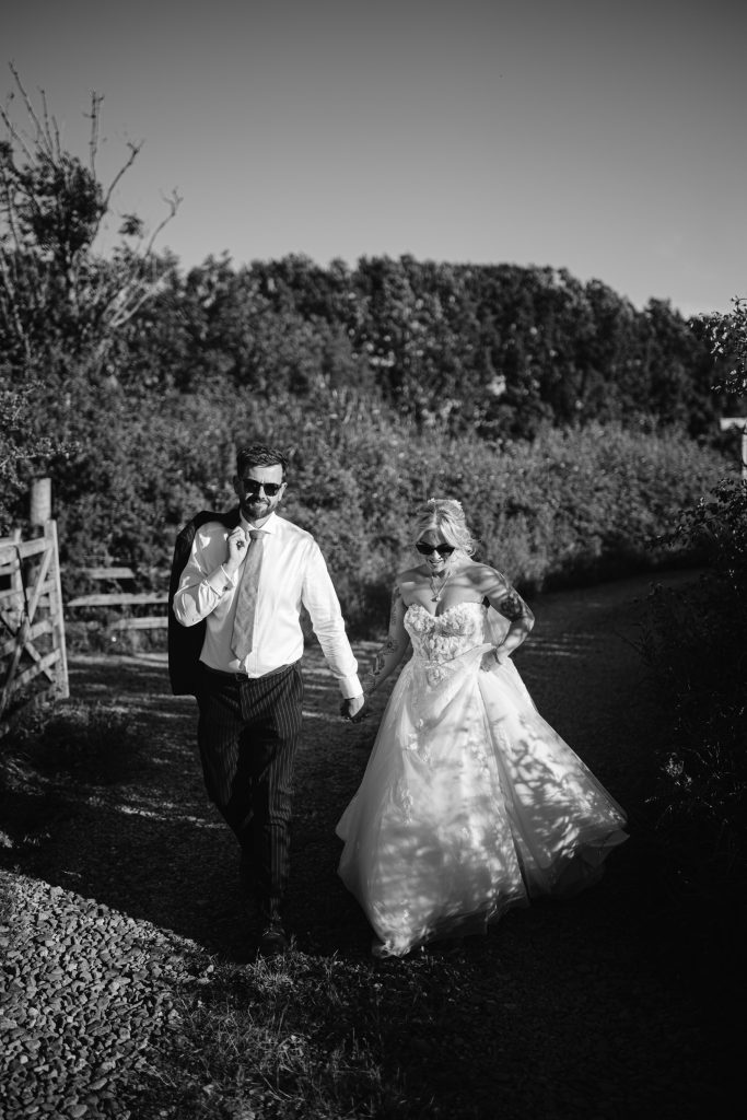 A man in a shirt and tie walks beside a woman in a wedding dress along a gravel path, both wearing sunglasses, with trees and a wooden gate in the background.