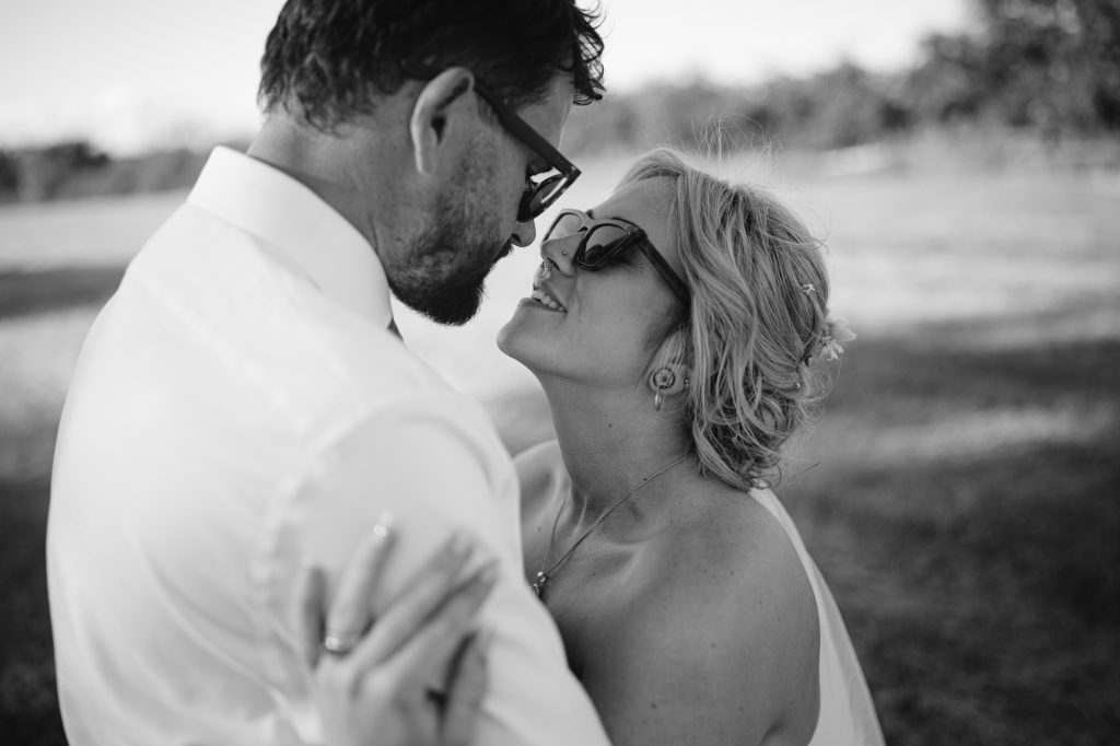 A man and woman wearing sunglasses embrace closely outdoors, looking at each other and smiling.