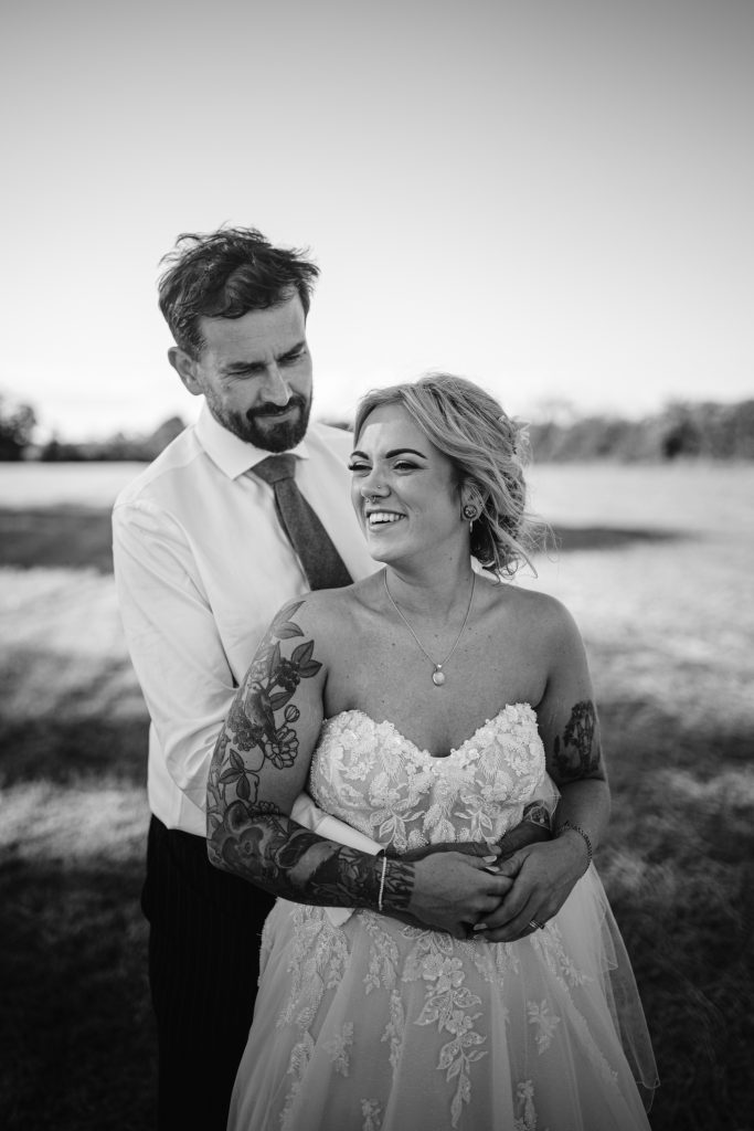 A man in a shirt and tie stands behind a woman in a lace wedding dress, both smiling, in an outdoor grassy field.