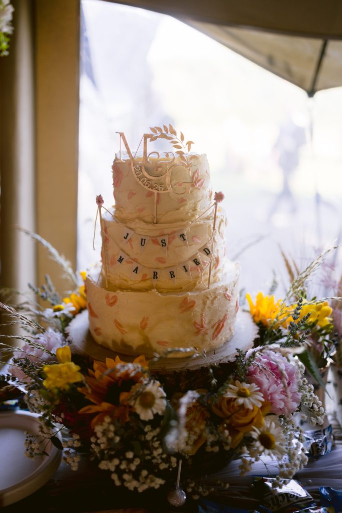 Three-tier wedding cake with floral decorations, a "JUST MARRIED" banner, and a "Mr & Mrs" topper, surrounded by an arrangement of assorted flowers.