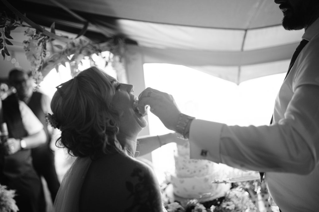 A bride sits and opens her mouth as a groom feeds her cake at a wedding reception; a decorated cake is visible in the background.