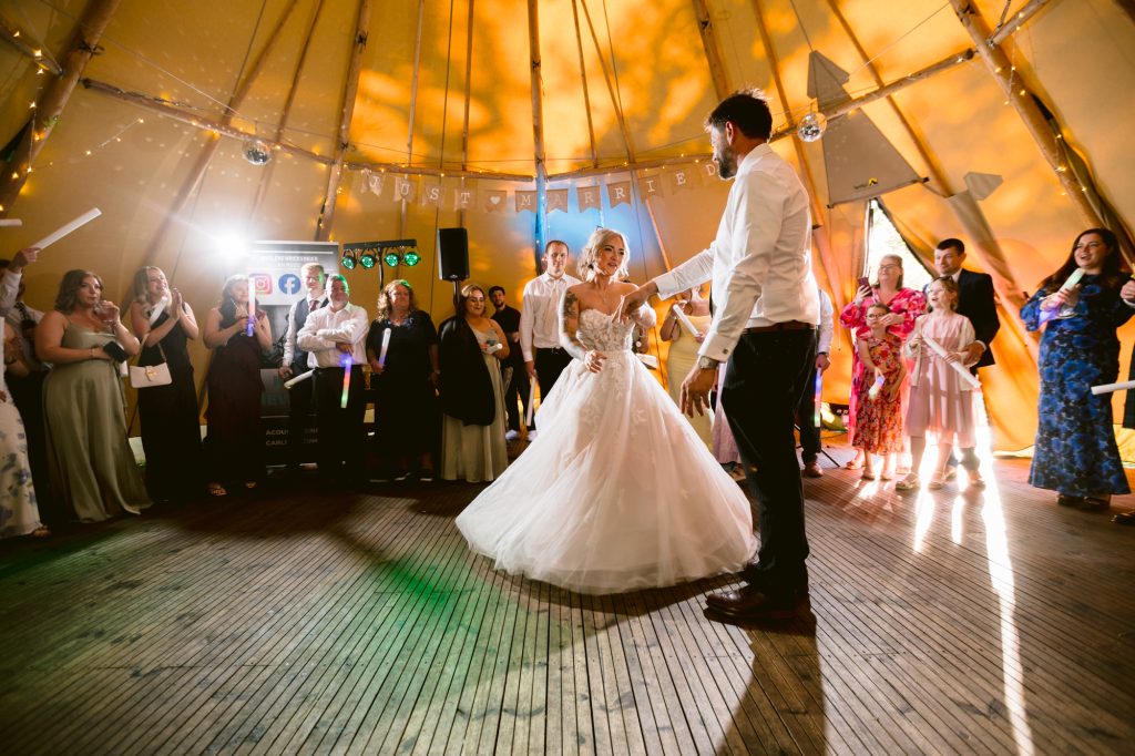 A bride and groom dance in the centre of a wooden floor surrounded by guests inside a decorated marquee.