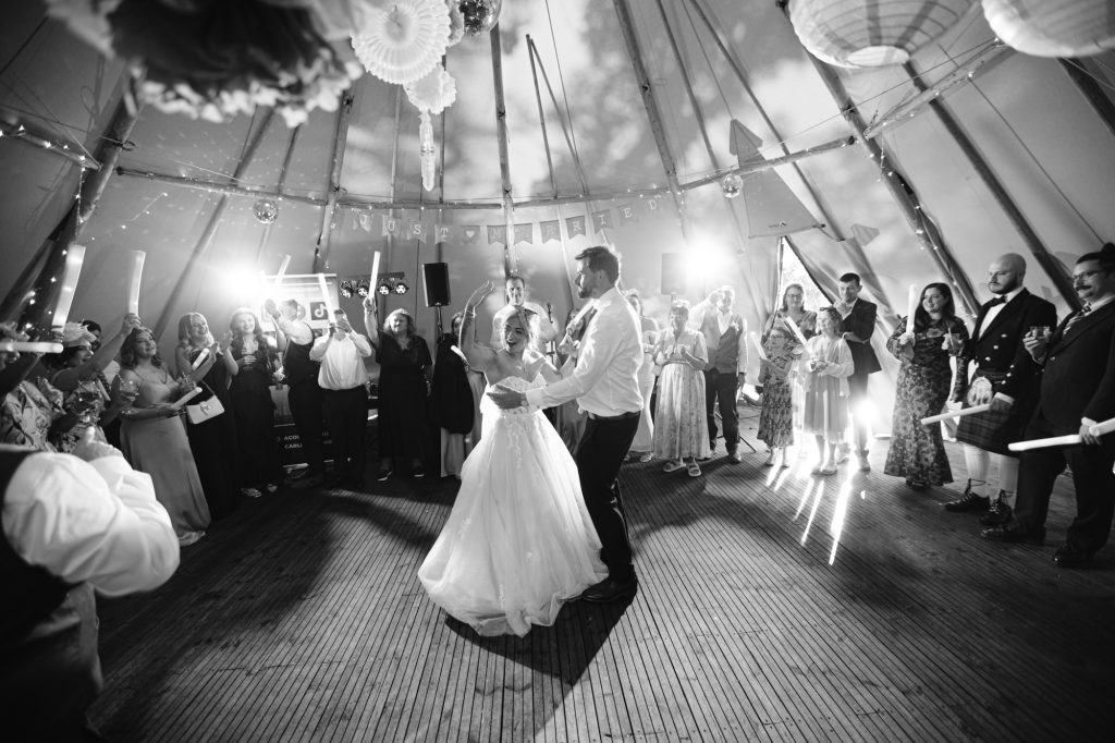 A bride and groom have their first dance inside a marquee, surrounded by guests watching and taking photos.