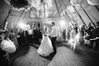 A bride and groom have their first dance inside a marquee, surrounded by guests watching and taking photos.