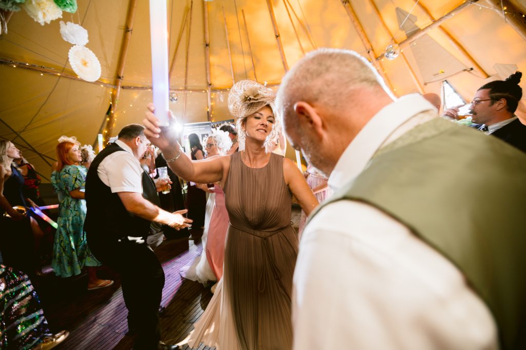 Woman in a taupe dress and decorative hat dances with a man in a waistcoat at a lively event inside a marquee decorated with lights and paper flowers.