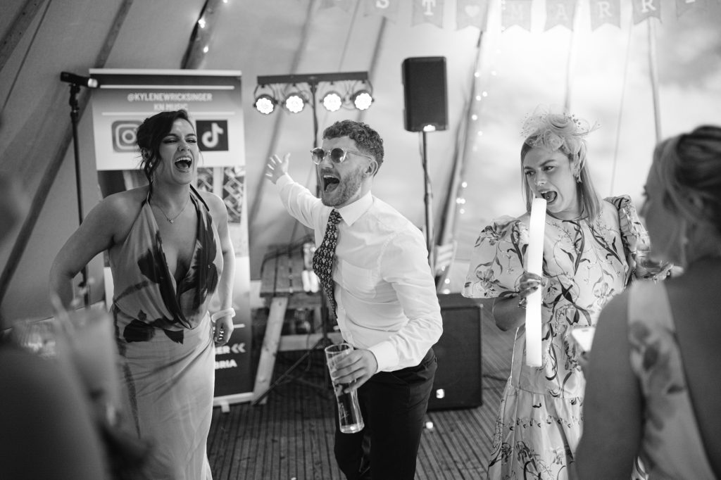 Three people sing and dance enthusiastically at a party, holding drinks and glow sticks, with lights and a banner in the background.