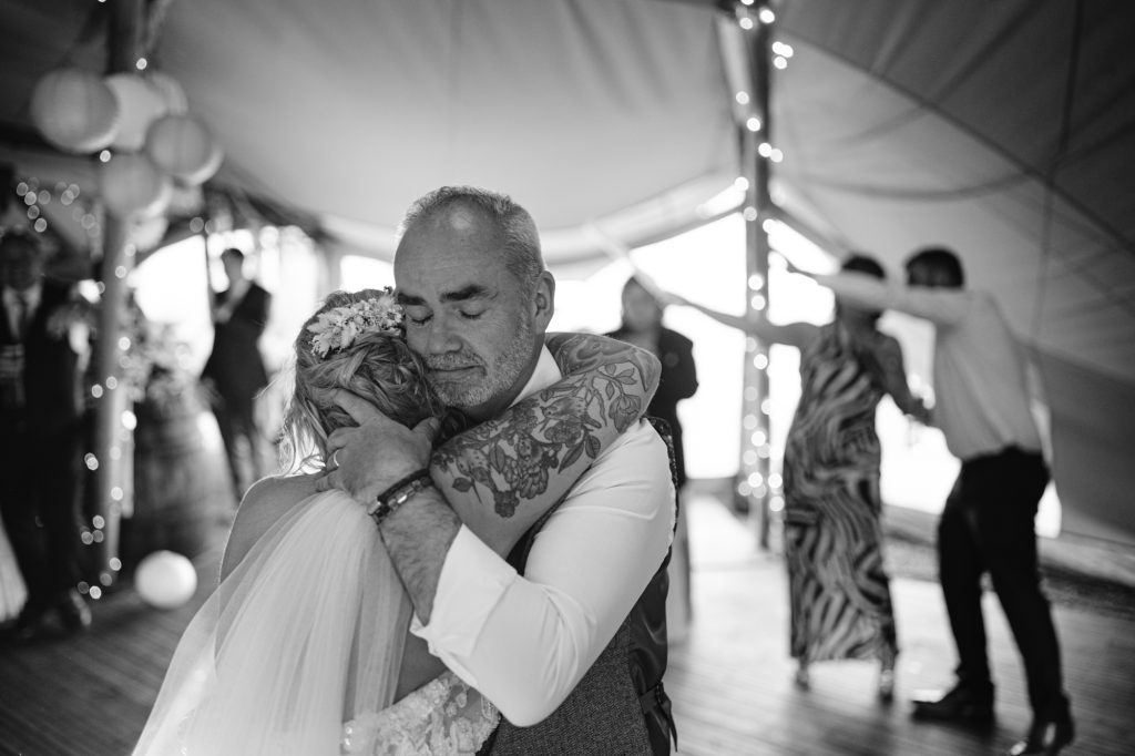 A bride and an older man embrace closely on a dance floor at a wedding reception, with other guests dancing in the background.