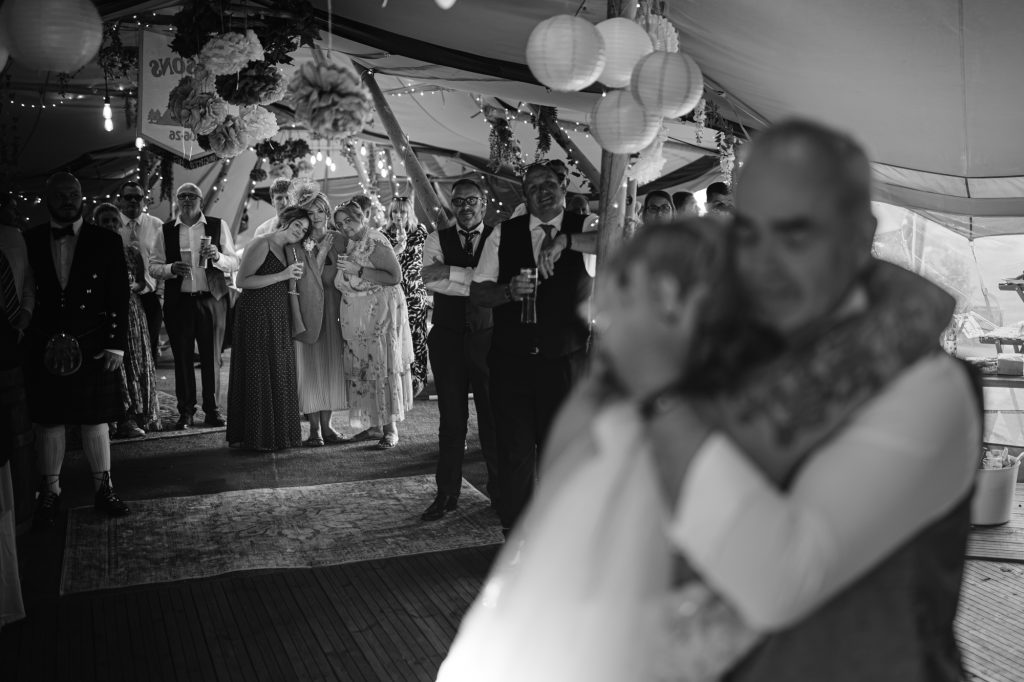 A black and white photo shows guests watching a couple embracing and dancing at a wedding reception under a decorated marquee.