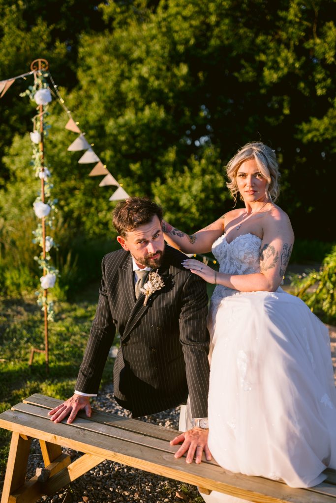 A bride in a white dress poses playfully behind a groom in a striped suit, both outdoors by a wooden bench with greenery and wedding decorations in the background.