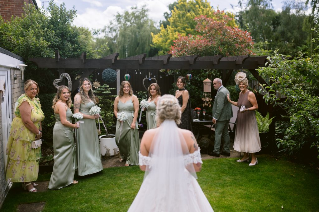 A bride in a white dress stands facing six bridesmaids in green dresses and two adults in a garden, preparing for a wedding ceremony.