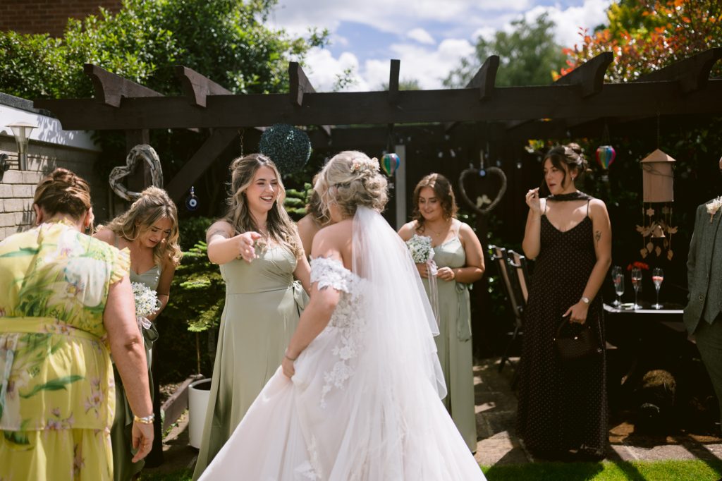 A bride in a white dress is surrounded by bridesmaids in sage green dresses and a few guests at an outdoor wedding reception.