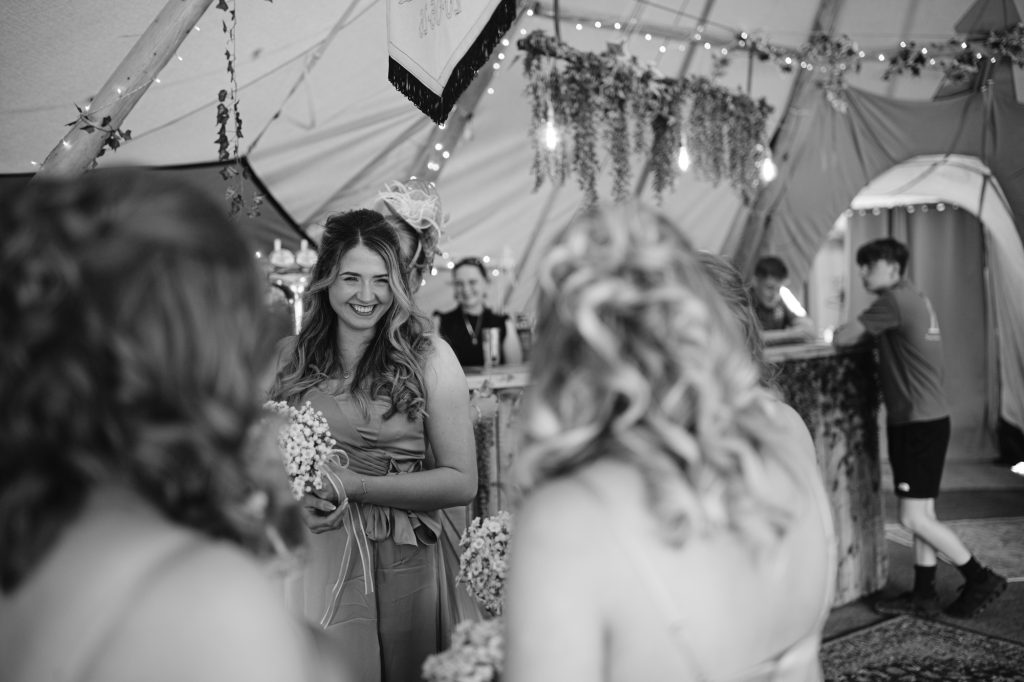 Three women in dresses stand together indoors, one smiling and holding flowers; bar area and fairy lights visible in the background. Black and white photo.