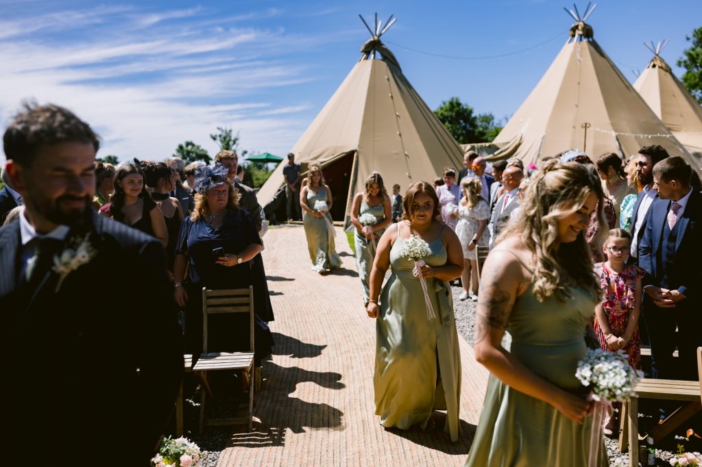 Bridesmaids in green dresses walk down an outdoor aisle lined with guests, with large beige tipis set up in the background under a blue sky.