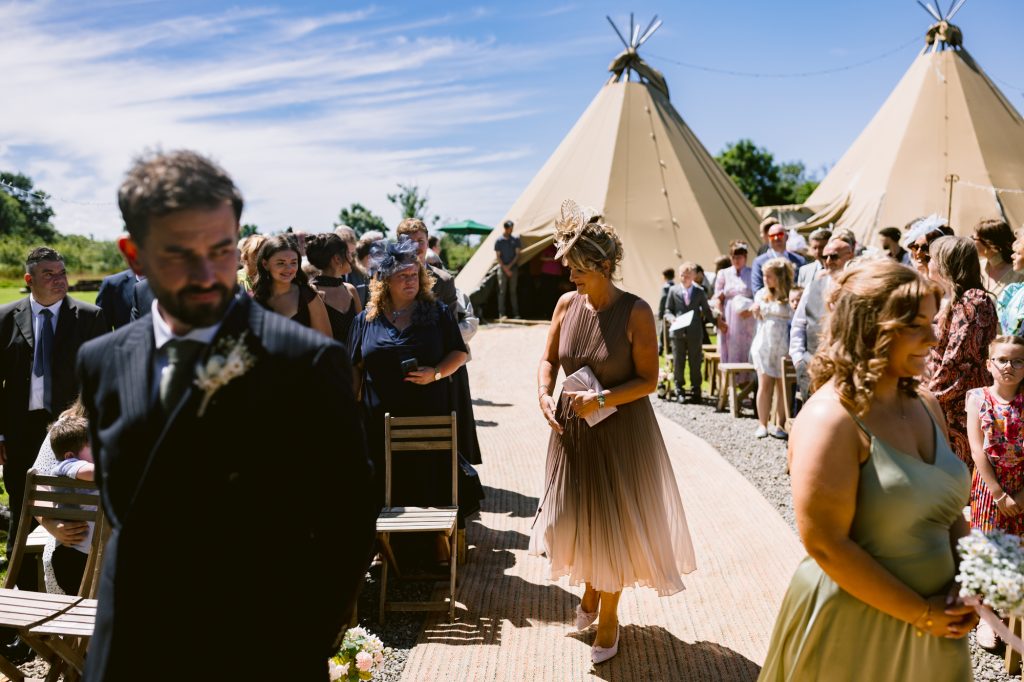 Guests walk down an outdoor aisle towards tipi tents under a blue sky, with people seated and standing on either side, at a wedding ceremony.