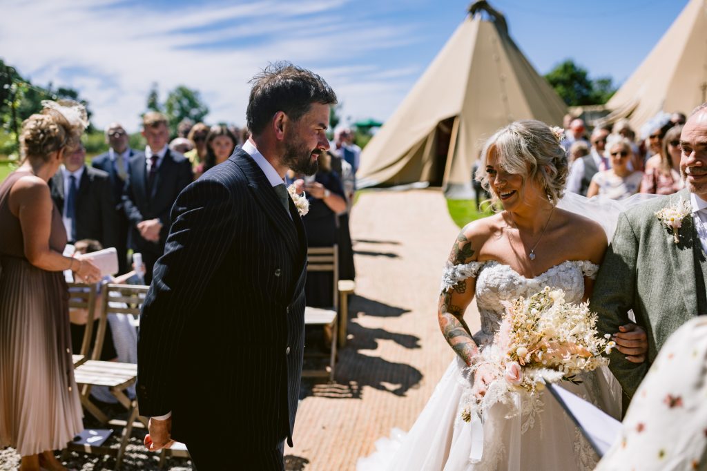 A bride and groom smile at each other during an outdoor wedding ceremony, with guests seated around them and large marquees in the background.