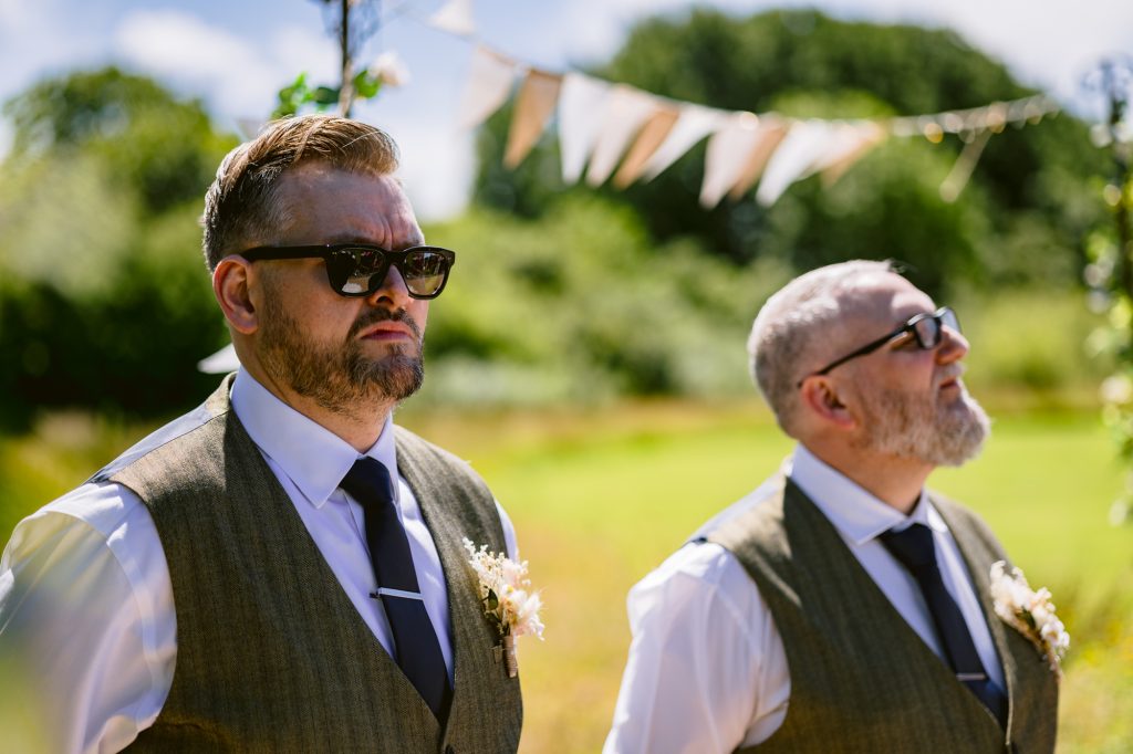 Two men wearing sunglasses, white shirts, ties, and waistcoats stand outdoors at a sunny event, with greenery and bunting visible in the background.