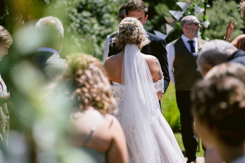A bride in a white gown and veil stands facing a groom during an outdoor wedding ceremony, surrounded by guests and greenery.