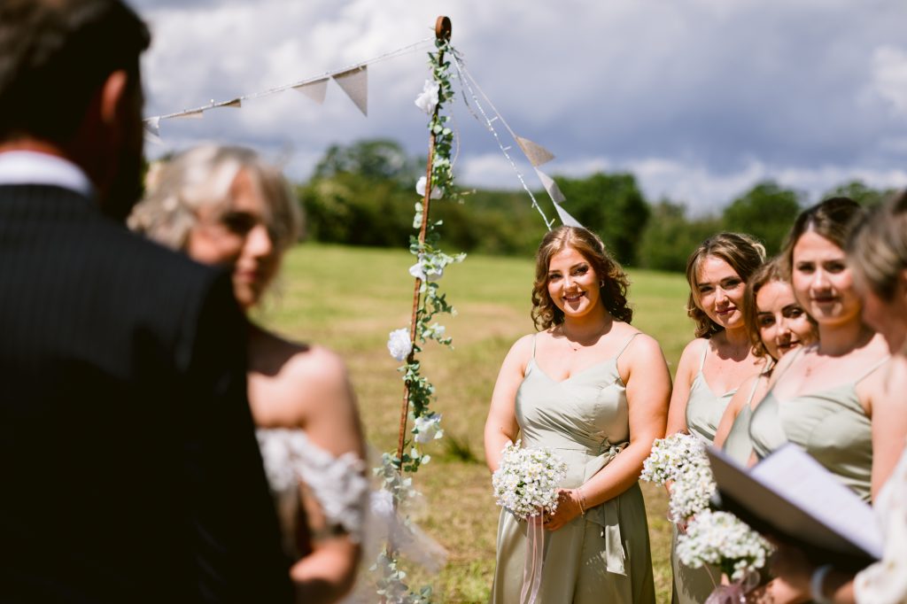 A group of bridesmaids in light green dresses stand outdoors, holding flowers, during a wedding ceremony with greenery and bunting decorations.