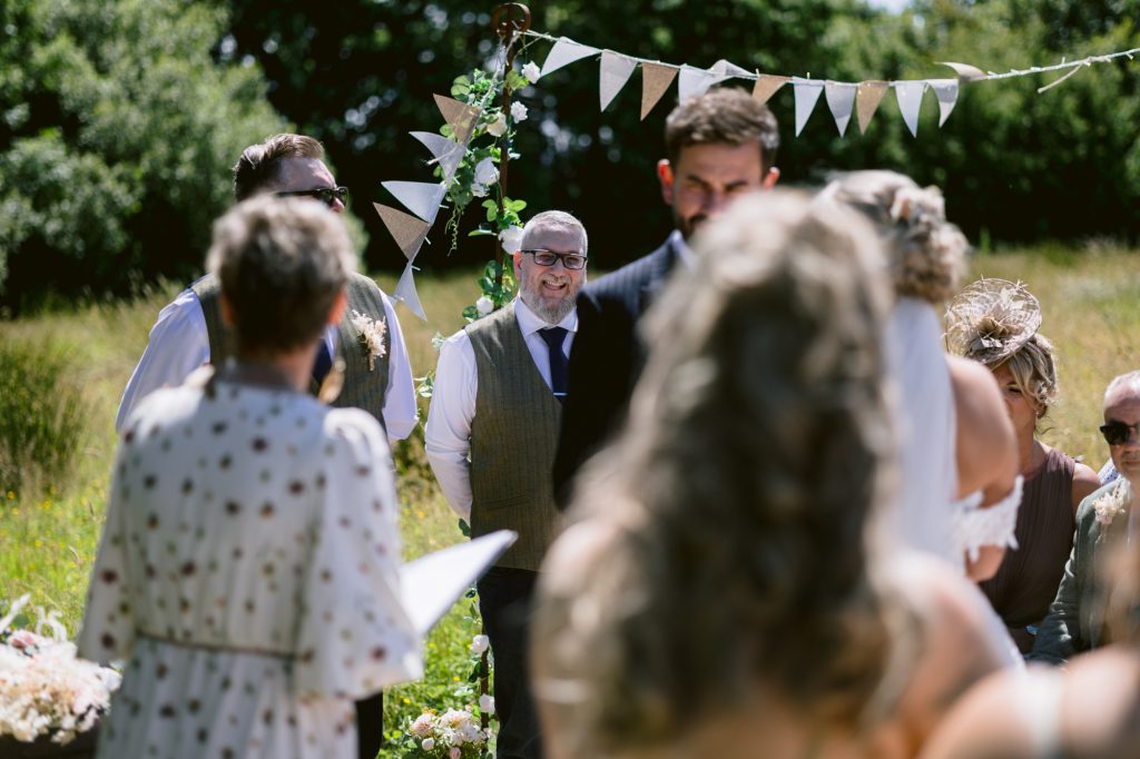 An outdoor wedding ceremony with guests seated and a man in a waistcoat and tie standing under bunting decorations, surrounded by greenery.