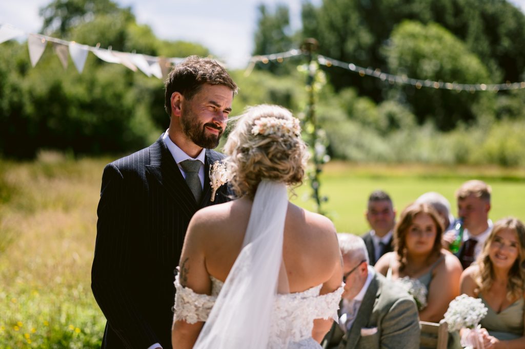 A bride and groom stand facing each other outdoors during their wedding ceremony, with guests seated behind them and greenery in the background.