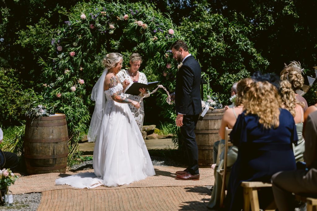 A bride and groom stand facing each other during an outdoor wedding ceremony, with an officiant reading and guests seated nearby. A floral arch and greenery are in the background.