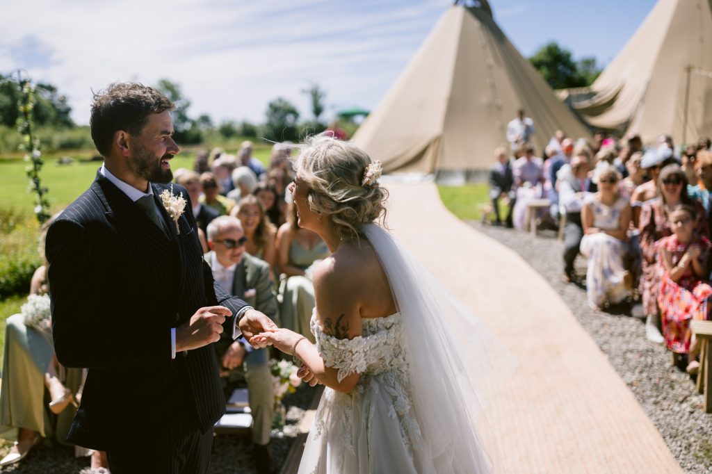 A bride and groom exchange rings during an outdoor wedding ceremony with guests seated in the background and large marquees behind them.