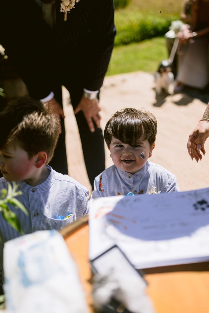 Two young boys in striped shirts stand outdoors near a table with papers. One boy is smiling at the camera whilst an adult stands behind them.
