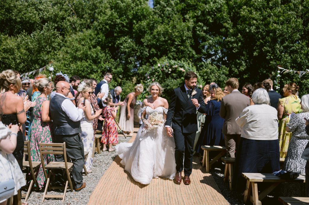 A bride and groom walk down the aisle outdoors as guests seated and standing on both sides clap and celebrate their wedding. Trees and sunlight are visible in the background.