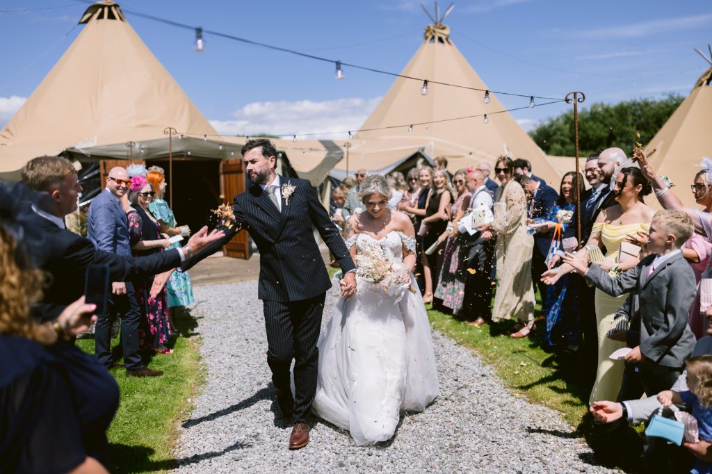 A bride and groom walk hand-in-hand down an outdoor aisle, surrounded by guests celebrating with confetti near tipi tents under a sunny sky.