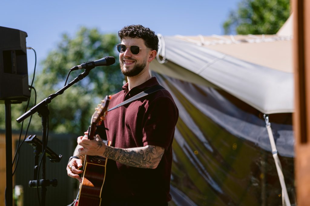 A man with sunglasses plays an acoustic guitar and sings into a microphone outdoors, standing near a tent on a sunny day.