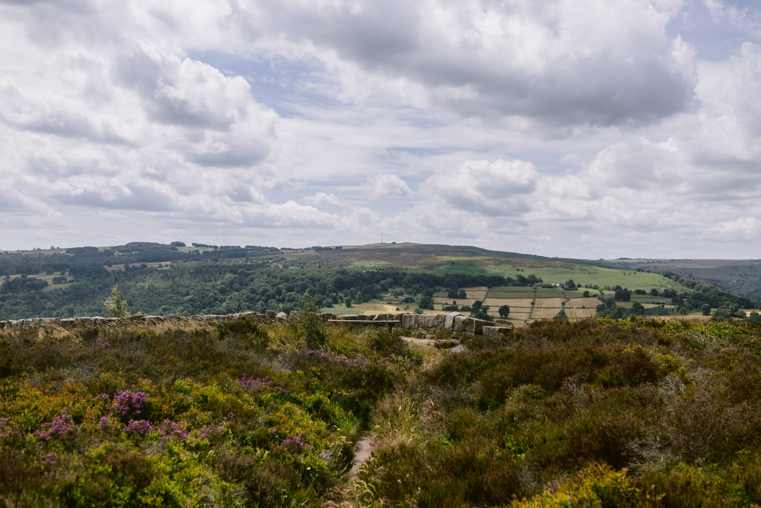 A cloudy sky drapes over the rural Peak District landscape, with green fields, distant hills, and patches of wildflowers creating a picturesque scene perfect for a proposal near Surprise View.