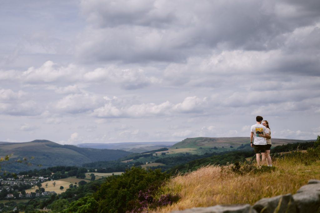 Two people stand on a grassy hilltop at Surprise View in the Peak District, overlooking a valley with distant hills under a cloudy sky—an unforgettable spot for a proposal.