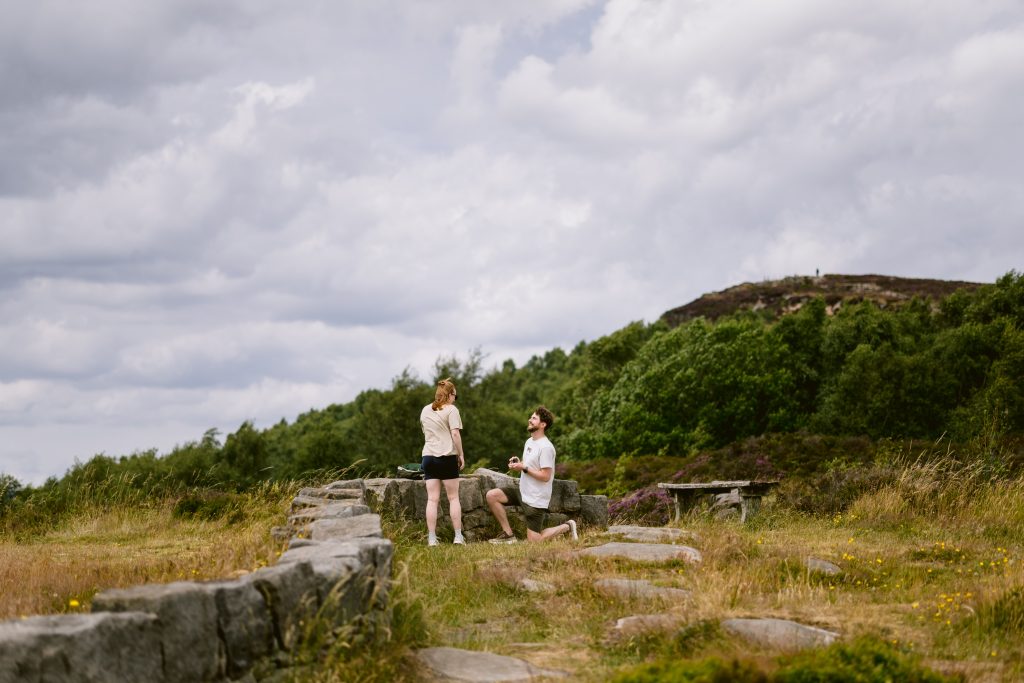 A man kneels on one knee holding a ring, proposing to a woman on a grassy path with stone steps and trees at Surprise View in the Peak District.