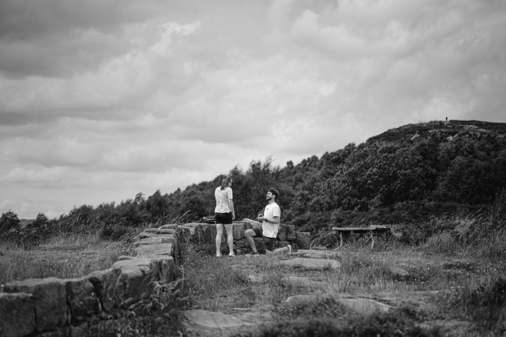 A person kneels and proposes to another on a rocky path at Surprise View in the Peak District, with rolling hills and cloudy skies in the background.