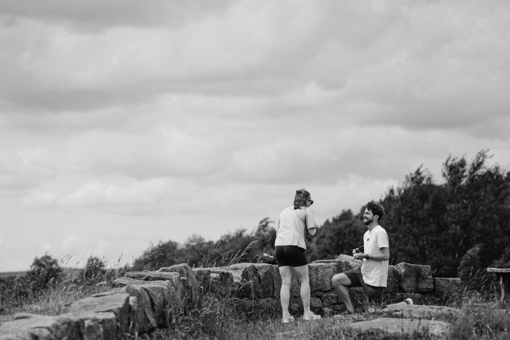 A man kneels on one knee in front of a woman outdoors near a stone wall at Surprise View in the Peak District, suggesting a heartfelt marriage proposal.