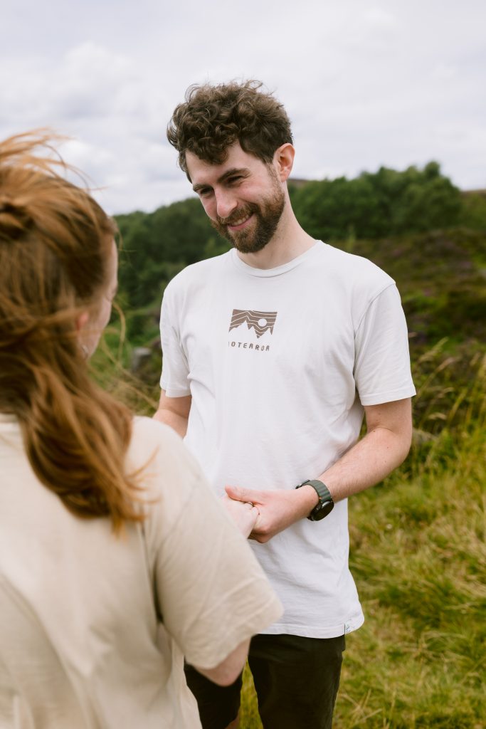 A man and woman stand outdoors at Surprise View in the Peak District, facing each other and holding hands, with greenery and a cloudy sky in the background—capturing a perfect proposal moment.