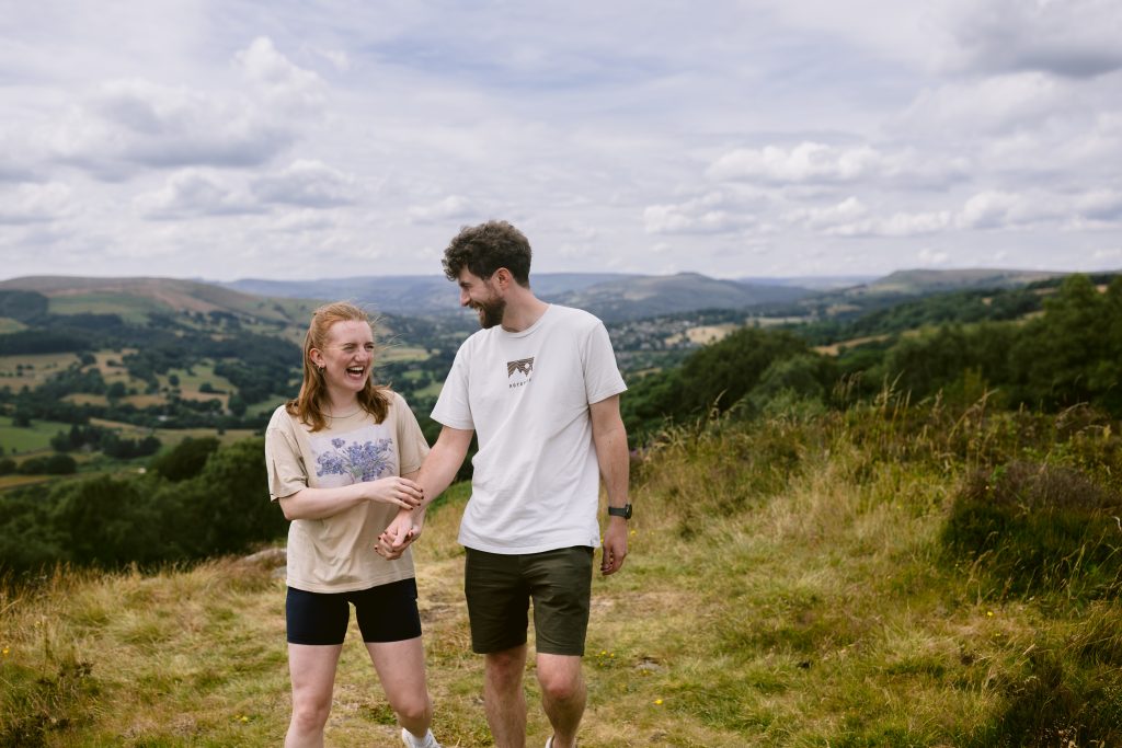 Two people walk outdoors on a grassy hill in the Peak District, smiling and holding hands, with rolling hills and a cloudy sky in the background.
