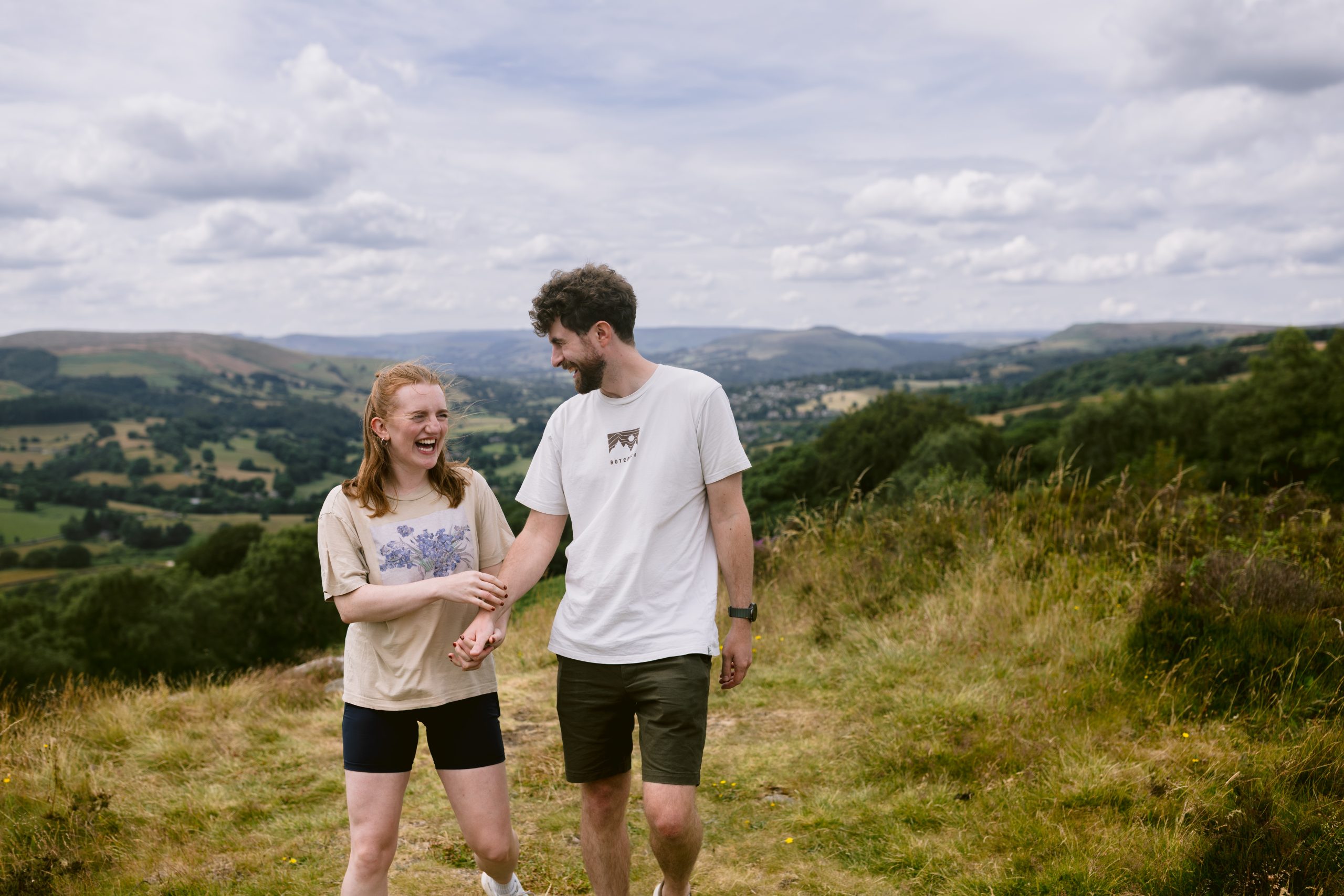 Two people walk outdoors on a grassy hill in the Peak District, smiling and holding hands, with rolling hills and a cloudy sky in the background.
