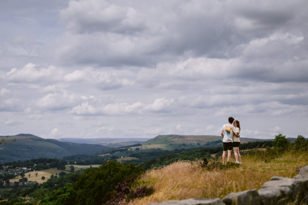 Two people stand together on a grassy hill in the Peak District, overlooking a scenic landscape of rolling hills and cloudy sky—an unforgettable spot for a proposal.