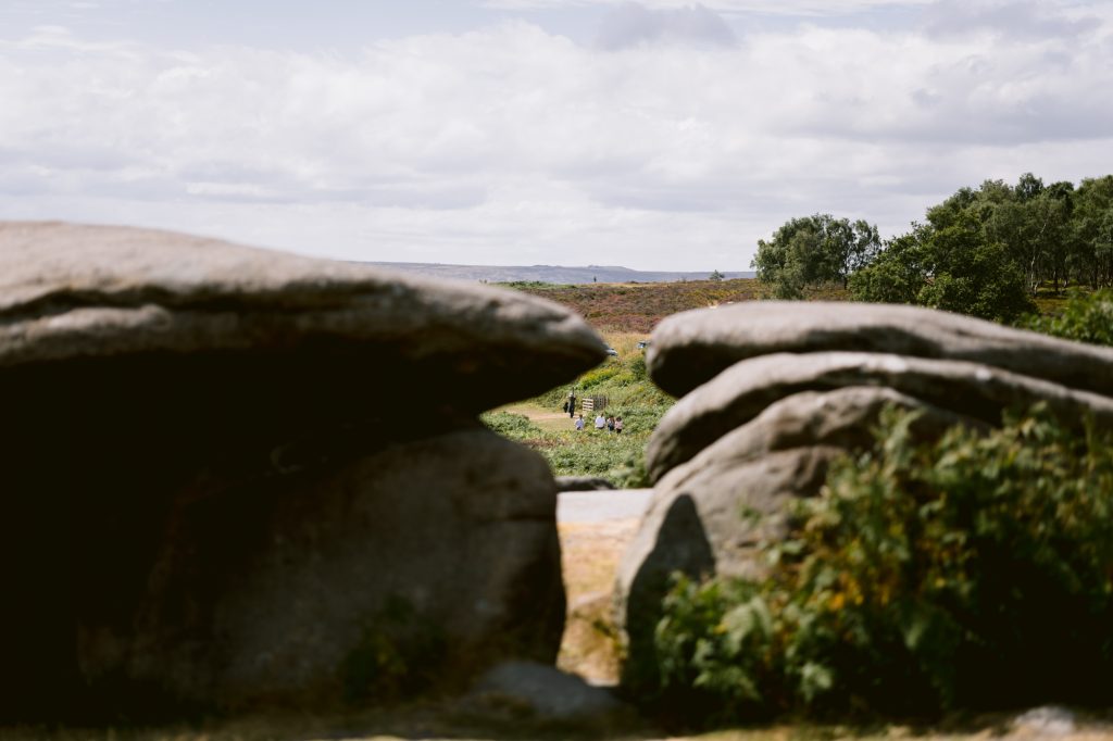 Large rocks frame a distant view of people walking along a green path under a partly cloudy sky in a picturesque Peak District countryside setting near Owler Tor.