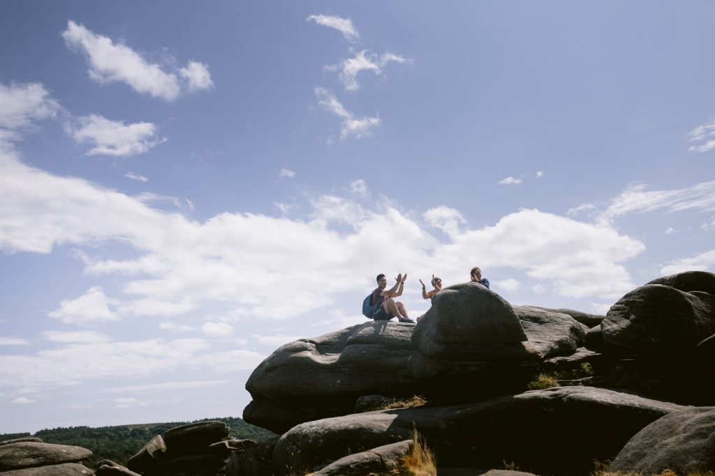 Three people sit on large rocks at Owler Tor under a blue sky with scattered clouds, enjoying the stunning Peak District view and gesturing with their hands—perhaps sharing stories or celebrating a surprise engagement.