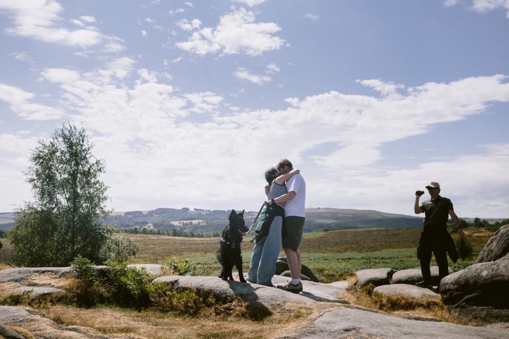 Two people share a heartfelt hug on the rocky landscape of Owler Tor in the Peak District, with a dog beside them and another person standing nearby, capturing the special moment—perhaps even a surprise proposal—against a backdrop of partly cloudy skies and open countryside.