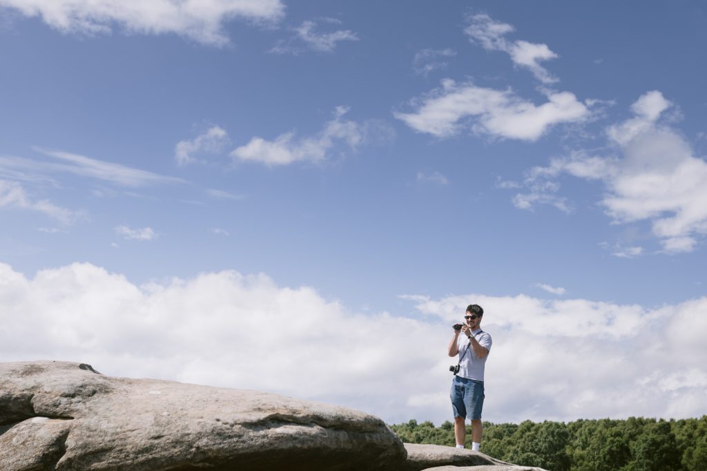 A man wearing sunglasses stands on a large rock at Owler Tor in the Peak District, taking a photo with a camera under a blue sky with scattered clouds.