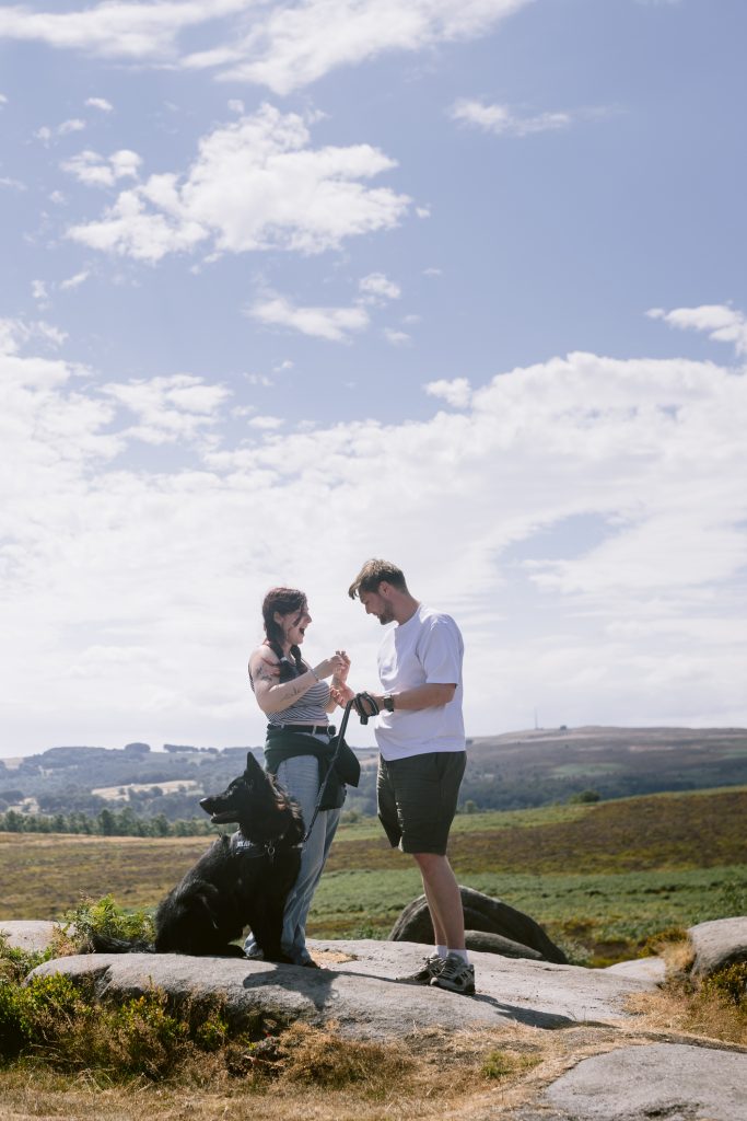 Two people stand on rocks outdoors with a black dog sitting beside them at Owler Tor in the Peak District. The landscape features grassy fields, distant hills, and a partly cloudy sky—creating the perfect backdrop for a surprise proposal.