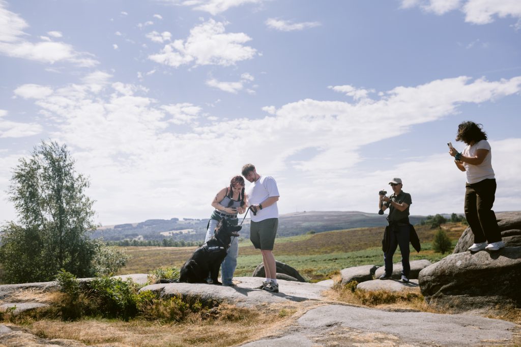 Four people with cameras and a black dog stand on rocky terrain at Owler Tor in the Peak District under a partly cloudy sky, with open countryside and hills in the background—an idyllic setting for a surprise proposal.